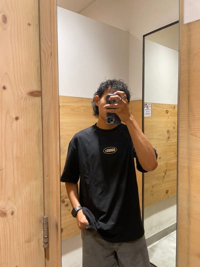A person stands in a changing room, taking a mirror selfie. The person is wearing a black t-shirt with a white oval logo that reads "2000s" and gray pants. They have curly dark hair and are wearing rings on their fingers and a watch on their left wrist. The changing room has light-colored wooden walls and a white ceiling. The floor is tiled. A partially visible sign is attached to the wall to the right of the mirror, which is a reflective surface. The overall environment suggests a retail or fitting room space. The person is in Kelapa Dua, Indonesia.