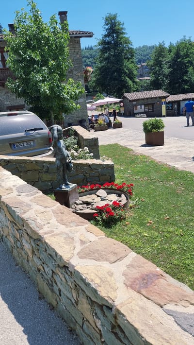 The image depicts a sunlit outdoor scene in Rupit, Spain. In the foreground, a stone wall runs diagonally across the frame. To the right of the wall is a green lawn with scattered dry leaves. A small circular fountain with rocks and red flowers is visible. A bronze statue of a child holding buckets stands on a pedestal in front of the fountain. Behind the statue, a light-colored SUV with a license plate reading "8242 DZL" is parked. In the background, several stone buildings with tiled roofs are present, some with awnings providing shade. People are seated at tables under a pink umbrella. Further back, more buildings and lush green trees are seen, with hills in the distance under a clear blue sky.