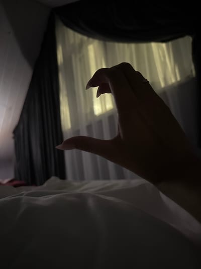 A close-up, low-light, over-the-shoulder shot shows a left hand forming a heart shape against a backdrop of sheer white curtains illuminated by a warm, yellow light. The hand, with long, manicured fingernails, is positioned in the foreground and casts a shadow on the white, rumpled sheets of a bed. Dark, heavy curtains frame the window, creating a somber atmosphere. The location is identified as Doma 1096 Km, Russia.