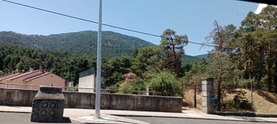 The image depicts a residential area in San Rafael, Spain, characterized by a mix of houses and natural vegetation. In the foreground, a concrete barrier runs horizontally, topped with a grey metal railing. To the left, a weathered concrete object with circular indentations sits on the pavement. A tall, grey metal pole stands prominently in the mid-ground, near the center of the frame. Behind the barrier and pole, several residential buildings with terracotta roofs are visible. Further back, a lush, green hillside covered in dense pine forest rises towards a clear blue sky. The overall scene suggests a sunny day in a semi-mountainous region.