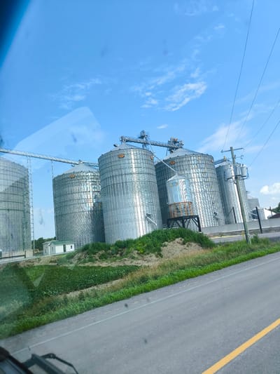 In North Stormont, Canada, a group of large, corrugated metal grain silos stands under a clear blue sky with scattered clouds. The silos are interconnected by elevated conveyor systems, suggesting an active agricultural or industrial facility. A small, white building with a green roof is situated near the base of the silos on a sloped, grassy embankment. Power lines and a utility pole are visible to the right of the silos. In the foreground, a paved road with a yellow dividing line curves into the frame, and the edge of a vehicle's windshield wiper is visible at the bottom left, indicating the photo was taken from inside a moving vehicle. The overall environment appears to be rural, with a focus on agricultural infrastructure.