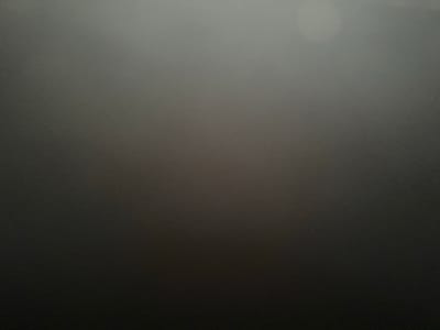 This is a photograph with a dark and abstract aesthetic, likely taken with a camera that had difficulty focusing or was in low-light conditions. The image is predominantly dark gray, with a gradient effect that transitions from a very dark, almost black hue at the top to a lighter, desaturated gray towards the bottom. There are no discernible subjects, objects, or clear scene details. The texture of the image is grainy, suggesting a photograph taken in low light or with a high ISO setting. The bottom of the image shows a slight blur, potentially indicating a lens flare or a very out-of-focus element. No text is visible. The overall impression is one of darkness and ambiguity, with no specific activity, event, time of day, weather, or location cues identifiable.