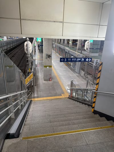 The image captures a downward view of a concrete staircase at a subway station in Uijeongbu-si, South Korea. The staircase is lined with metal handrails on both sides and has yellow tactile paving strips on some of the steps. At the bottom of the staircase, there is a covered section with a gray and black ramp-like structure.
At the top of the staircase, a subway platform is visible. To the left, train tracks run along the platform. To the right, a concrete pillar with black and yellow hazard stripes is present, along with a red fire extinguisher. A blue directional sign indicates "← 3 Yeomcheon Dongducheon Soyosan 4 →". Further down the platform, a green emergency exit sign with a running figure is illuminated. Several subway train cars are parked at the platform, with their doors closed. A few individuals can be seen on the platform, one standing near the emergency exit and another further down. The environment appears to be an indoor subway station, with overhead lighting and a somewhat industrial aesthetic.
