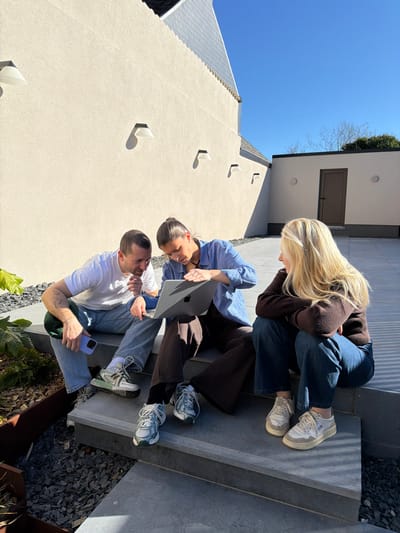 The image displays an outdoor courtyard scene in Marche-lez-Écaussinnes, Belgium, under clear blue skies. Three individuals are seated on dark grey stone steps, engaged with a silver Apple MacBook laptop.

On the left, an adult male with short hair and a white t-shirt, light blue jeans, and light-colored sneakers, holds a light purple-cased smartphone in his left hand. A tattoo is visible on his left forearm. He looks towards the laptop held by the central figure.
In the center, an adult female with dark hair tied back wears a blue and white checkered long-sleeved shirt over a dark top, dark wide-leg trousers, and light-colored sneakers. She holds the open laptop on her lap, actively manipulating the keyboard with her left hand while looking at the screen.
On the right, an adult female with long blonde hair, dressed in a dark brown long-sleeved top, dark blue jeans, and light-colored sneakers, observes the laptop and the central female. She is seated one step higher than the other two.

The background features a light beige, textured building wall with multiple white, angular wall-mounted light fixtures casting distinct shadows. A dark grey tiled roof is visible above this wall. Further right, a smaller, light-colored structure with a dark brown door and frame stands under the direct sunlight. Dark decorative gravel fills planting beds adjacent to the steps, containing minimal green foliage. The paving consists of dark grey stone slabs and steps.