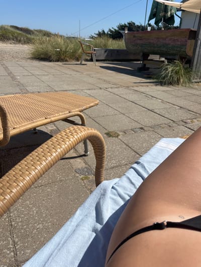 This image depicts an outdoor leisure scene under clear, bright sunlight, situated on the coast in Skagen, Denmark. The perspective is from a person reclining on a light brown wicker-style outdoor lounge chair, which is covered by a light blue and white striped towel. In the immediate foreground, the person's tanned upper thigh and hip are visible, clad in black bikini bottoms that feature a small silver connector ring on the hip strap. A small, dark tattoo is discernible on the upper right hip.

Adjacent to the lounge chair, a matching light brown wicker-style outdoor table or low bench is partially visible. The area around the seating is paved with light grey square concrete pavers, accented by darker brick lines. Strong sunlight casts distinct shadows across this paved surface.

Beyond the paved area, the environment transitions to sandy ground interspersed with tall, dry coastal grasses, indicating a dune-like landscape. A single light brown outdoor chair with a metal frame is positioned on this sandy section. Further into the background, a weathered wooden boat, painted with faded horizontal stripes of green, red, and yellow, rests elevated on wooden supports. Above and behind the boat, a green canopy or umbrella is partially visible, next to a white building or structure. The sky above is clear and blue.