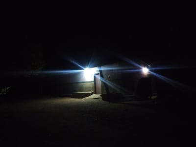 The image depicts a nighttime scene in Bajoga, Nigeria, at coordinates 10.852° N, 11.426° E. A small, single-story building is illuminated by two bright lights, casting long, distinct beams across the dark, dusty ground. The structure appears to have an open doorway and a small step leading up to it. A shadowy figure stands near the entrance, partially obscured by the light. The surrounding area is completely dark, suggesting a rural location with minimal artificial light. The image captures a simple, everyday scene in a dimly lit neighborhood.