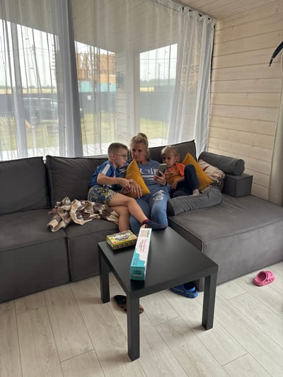 The image depicts a modern, light-colored wooden interior with large windows covered by sheer white curtains. A dark gray sectional sofa occupies the center of the room, with a black coffee table placed in front of it. On the sofa, a woman is seated between two children. The woman, with blonde hair pulled back, is wearing a blue sweatshirt and blue pants. She is holding a yellow pillow and looking at a smartphone in her hand. To her left, a boy in a blue and white t-shirt and patterned shorts sits with his legs crossed, also looking at the woman. To her right, a young girl with blonde hair is wearing an orange t-shirt and dark pants, looking intently at a smartphone. A yellow pillow is behind her. On the coffee table are two board games and a phone. A patterned blanket is draped over the left side of the sofa. On the floor, a pair of blue flip-flops are tucked under the coffee table, and a pink sandal is to the right of the coffee table. The background visible through the windows suggests an outdoor scene with greenery and some construction. The overall impression is one of a casual, indoor family gathering in what appears to be a residential setting. The city of Starye Chernitsy, Russia, is mentioned as the location, implying this scene is within that region.