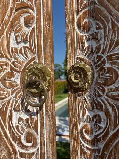 The image shows a close-up of a decorative wooden chest or cabinet door in Cemagi, Indonesia. The wood is light brown with white-washed carving details. Two bronze flower-shaped handles adorn the door, the lower one with a hanging ring. In the background, visible through the space between the doors, is a blue sky and some greenery, suggesting an outdoor setting.