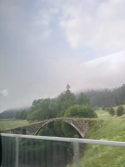 The image shows an old stone bridge spanning a body of water. The bridge has a single arch and appears to be made of rough-hewn stones. It is surrounded by lush green vegetation, including trees and grass. In the background, a dense forest stretches across rolling hills, partially obscured by a layer of mist. The sky above is overcast with patches of white clouds. The scene is serene and appears to be a natural landscape. Based on the provided context, this location is identified as the "Devil's Bridge" in the Arda River valley near Zmeitsa, Bulgaria. There are no people or animals visible in the image.