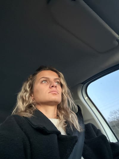 A woman with blonde, wavy hair and green eyes sits in a car. She wears a black coat over a white sweater. The car's interior is visible, including the sun visor and the sunroof, through which a bright, pale sky is seen. Her face is tilted upward, and she gazes off-camera.