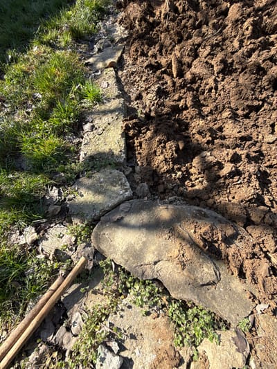 An overhead view depicts an outdoor ground-level scene in Devetaki, Bulgaria, characterized by a distinct division between a vegetated area and disturbed soil. The left side of the frame features a patch of green grass intermingled with dry leaves and small ground cover plants, illuminated by direct sunlight. A linear border of weathered, light grey concrete or stone slabs separates this grassy section from the adjacent area. The right side of the frame consists entirely of dark brown, freshly disturbed soil piled irregularly, indicating recent excavation or earth-moving. In the foreground, at the lower center-right, a large, irregularly shaped, flat grey stone or slab rests partially embedded in the ground, bridging the transition between the grassy area and the disturbed soil. Two light brown, slender wooden sticks or dowels lie parallel to each other on the ground in the bottom left corner. Shadows are cast by the concrete slabs and the large stone. No individuals or active interactions are present.
