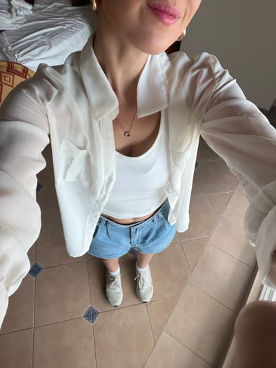 A single individual stands in an interior setting, likely a room in Chamilpa, Mexico, capturing a high-angle, downward-facing image of their attire. The person wears a white, ribbed tank top under an unbuttoned, sheer white long-sleeved shirt with a collar and a single left breast pocket. Light blue pleated denim shorts are worn on the lower body, exposing a portion of the abdomen. White socks and light-colored athletic shoes complete the ensemble. A gold-toned necklace with a small, circular, possibly jewel-encrusted pendant is visible on the chest, along with a gold hoop earring on the right side of the head. The subject's lips are coated with pink lipstick. The flooring consists of large, light brown square tiles interspersed with smaller dark blue and white patterned accent tiles at regular intervals. In the background to the left, a rumpled white bed with patterned bedding and a partial headboard is visible, indicating a bedroom.