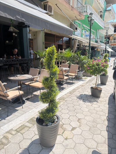 This is a medium shot, taken outdoors on a sunny day, looking down a pedestrian sidewalk lined with buildings and outdoor seating areas. The sidewalk is paved with light-colored hexagonal tiles.
In the foreground, a large, dark gray planter holds a manicured, conical evergreen tree. Further back, another similar planter holds a blooming pink bougainvillea tree. Several outdoor dining tables and chairs, upholstered in a light beige fabric, are arranged on the sidewalk.
On the left side of the image, the facade of a building is visible, featuring a green painted section with a sign that has blue and white text, though it's difficult to read. An air conditioning unit is mounted on the building. Part of an awning extends over a lower level with glass doors and dark metal railings. A man with sunglasses and a black t-shirt is seated at a table in this lower area, looking down at his phone.
The scene appears to be in a European town or city, possibly in Greece given the location context. The bright sunlight casts distinct shadows, suggesting it is daytime. The overall atmosphere seems relaxed and casual, typical of a café or restaurant setting.
