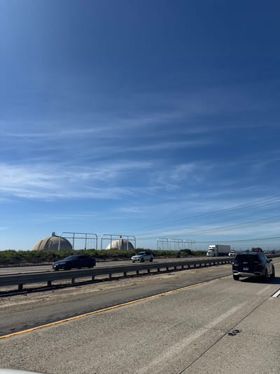 An elevated view alongside a highway displays clear blue skies with wispy clouds. Multiple vehicles, including a large semi-truck, are visible on the road. Power lines stretch across the top of the frame. Large, circular dome-like structures, possibly storage tanks, are situated near the roadside, separated by a guardrail. The highway pavement is light gray with marked lane divisions.