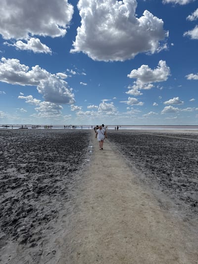 The image depicts a wide, flat landscape under a bright blue sky with scattered white clouds. A dirt path, slightly elevated and lighter in color, runs from the foreground towards the horizon. On either side of the path, the ground is dark and appears to be muddy or wet earth with visible indentations and textures. In the distance, a body of pinkish water stretches across the horizon, meeting the sky. A group of people are visible along the edge of the water and further out on the flat terrain. One person, wearing a white dress, is walking on the path in the middle ground, facing away from the camera. Another person, also in a white outfit, is walking on the path from the opposite direction, closer to the camera and to the left. The environment suggests a natural setting, possibly a salt lake or a mudflat. The location is identified as Бурсоль, Russia.