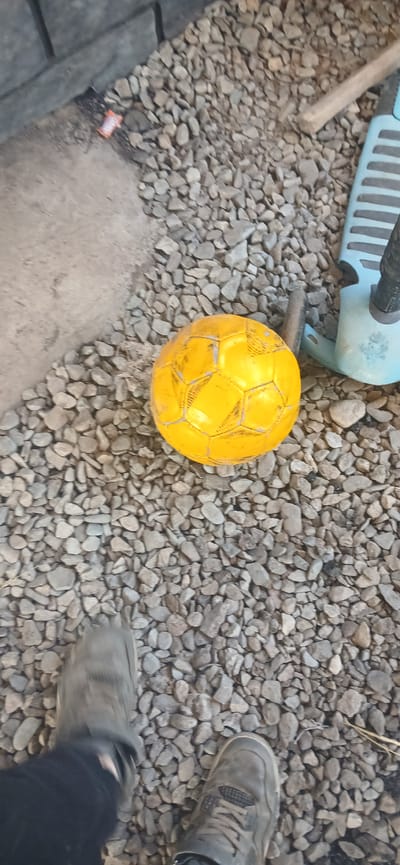 A high-angle outdoor shot shows a yellow soccer ball sitting on a gravel surface. To the right of the ball is a blue and black scooter. In the foreground, two feet are visible, one in a dark shoe and the other in a gray sneaker. The background features a concrete wall and more gravel. The scene is likely in Mihaylovka, Kyrgyzstan.