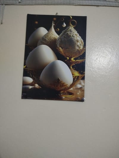 A poster hangs on a white wall, depicting four white eggs placed in golden bowls. The eggs are smooth and reflective, some more ornate than others. In the background, one egg has a pointed top with swirling patterns, while another is decorated with intricate details. Small beads surround the bowls, enhancing the image's luxurious and ornamental feel.