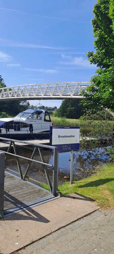 The image captures a sunny day scene at a waterway in Enniskillen, United Kingdom. In the foreground, a wooden walkway with a metal railing leads towards the water. A blue and white motorboat is docked at a pier. A sign with the text "Broadmeadow" and "Waterways Ireland" is prominently displayed next to the boat. Several ducks are swimming in the dark water. In the background, a white truss bridge spans the waterway, with lush green trees lining the banks. The sky is clear blue with some wispy clouds and a faint airplane contrail. The environment appears serene and recreational.