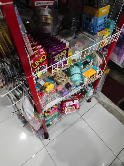 The image shows a close-up of a red metal shelf stocked with various toys and games. The shelf is set up in an indoor retail environment, possibly a small shop or stall. The items on display include board games like UNO, building block sets labeled "BRICKS," and smaller toy items such as toy cars and plastic figurines. Some of the items are still in their packaging. The lighting suggests an overhead light source. The floor visible at the bottom of the image is tiled with a light gray color. The overall impression is of a commercial display area focused on children's playthings. There are no people visible in the image. The specific location in Sidorejo, Indonesia, cannot be determined from the visual information provided in the image alone.