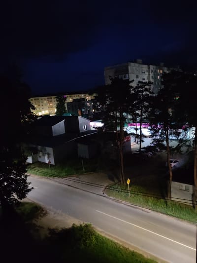The image depicts a night scene in the city of Kotovsk, Russia. The foreground shows a paved road curving to the left, bordered by grassy areas and trees. A few streetlights illuminate the road. In the middle ground, there are several low-rise buildings with dark roofs, appearing to be residential or commercial structures. Beyond these buildings, larger, multi-story apartment buildings are visible, their windows lit with warm light, indicating occupancy. Tall pine trees are scattered throughout the scene, their dark silhouettes contrasting with the illuminated buildings. The sky is a deep blue, transitioning to black at the top, with some faint cloud cover. Several cars are parked in a lot to the right of the middle-ground buildings. A yellow road sign is present on the right side of the road.