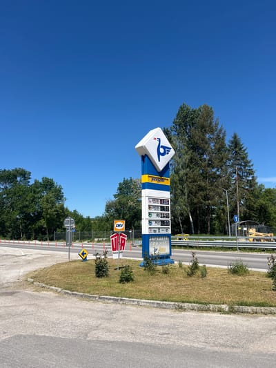 The image depicts a gas station in Malinovo, Bulgaria. A prominent sign displays fuel prices: Diesel at 1.86, Greenforce Diesel at 1.8, 95 octane fuel at 1.57, and Blue at 1.77. The sign also advertises restroom facilities and the gas station's branding. Below the gas station sign is a smaller sign with DKV on it. In the background, a road and trees are visible, as well as a yellow Gehl construction vehicle. The sky is clear and blue.