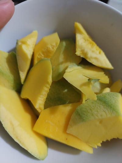 A close-up, overhead shot shows a white bowl filled with sliced mangoes. The mangoes are cut into irregular, wedge-shaped pieces, with some pieces displaying a yellowish-orange interior and others showing a greenish-yellow exterior. The bowl is white and appears to be ceramic. The lighting is warm and natural, casting soft shadows on the fruit. There is no indication of the specific location within the city of Jelutung, Indonesia.