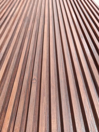 The image shows a close-up, angled view of a wooden surface composed of many parallel vertical planks. The wood has a rich, dark brown color with visible grain patterns and subtle variations in tone, suggesting it is a natural material, possibly teak or walnut. The planks are narrow and appear to be uniformly spaced, creating a ribbed or slatted effect. The lighting highlights the texture and depth of the material, with shadows cast in the grooves between the planks. There are no people, animals, or specific objects visible. The background is entirely the wooden surface, and there are no contextual elements to indicate a specific location within Settimo Torinese, Italy, or any other city. The image is abstract and focuses solely on the material and its pattern.