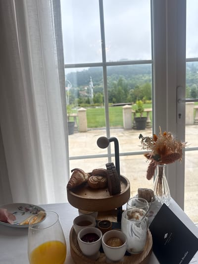 A breakfast tray is presented on a table in front of a window. The view from the window shows a landscape with a green field, trees, and what appears to be a distant building or structure. The location is Vila Verde, Portugal. On the tray, there is a variety of breakfast items including bread, pastries, yogurt in glasses, small cups with what appear to be spreads or jams, and a glass of orange juice. There is also a small vase with dried flowers and a menu card from MAISON ALBAR, Amoure. A curtain is partially visible to the left of the window.