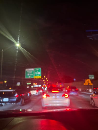 The image shows a view from inside a car, likely on a highway in North Miami, United States, at night. Traffic appears congested, evidenced by numerous red taillights and blurred headlights. Road signs are visible, including green exit signs, suggesting the presence of highway exits. The view is partially obscured by the car's windshield, which shows some light reflections and possibly raindrops. The scene depicts a typical nighttime traffic scenario.
