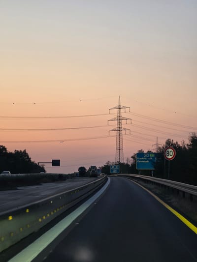 The image depicts a highway scene at dusk, likely on the A60 motorway near Bischofsheim, Germany, as indicated by a road sign. The sky is a gradient of soft orange and pale blue, suggesting the sun has recently set. A large electrical pylon with multiple power lines stretches across the upper portion of the frame. In the distance, other pylons and trees are visible. The road itself is dark asphalt, with a reflective yellow line on the right edge. A concrete barrier with yellow reflectors runs along the right side of the road.

On the road, traffic is light. A white car is visible in the far left lane, and another white car is further ahead. Construction vehicles, including a truck and a front loader, are parked on the right shoulder, indicating ongoing roadwork. A blue highway sign indicates directions to "Frankfurt a. M." and "Darmstadt," with distances of 35 km and 2 km respectively, and also shows "E42." Another sign on the left indicates "Rüsselsheim - Mitte 1000 m." A circular speed limit sign shows "80." The overall scene is calm and transitional, capturing the end of a day on a German autobahn.