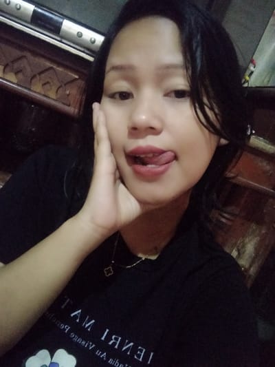 A young woman poses for a selfie. Her black hair is pulled back, and she wears a black t-shirt with white writing and floral details. She is sticking her tongue out and touching her cheek with her hand. A gold necklace with a clover pendant is visible. The background includes dark wood furniture, creating an indoor setting.