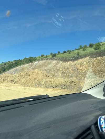 An image shows a view from inside a car, looking out through the windshield at a quarry. The quarry is a man-made excavation in the ground, exposing layers of rock and soil. The landscape is dominated by earthy tones, with brown and tan soil piles in the foreground and layered rock formations in the midground. The background features a green hillside covered in grass and scattered trees, with a clear blue sky above. There are some reflections visible on the windshield, including what appears to be a sticker or decal with circular markings. The dashboard of the car is visible in the lower part of the image, with a windshield wiper also visible. The scene depicts a construction or mining site, with signs of excavation and earth moving.