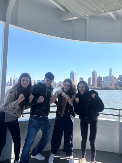 Four individuals pose for a photograph on what appears to be a boat or ferry. The backdrop features a cityscape with various buildings. The woman at the top is wearing a black fur-trimmed jacket and black pants. Below her, the second woman has long brown hair, sunglasses, a patterned Louis Vuitton-style scarf. The man in the middle wears a black jacket, glasses, and blue jeans. The woman at the bottom has long brown hair and is wearing a tan trench coat and black pants. All four individuals are clenching their fists and are posing to the camera. The shot is framed by what seems to be the metal frame of the boat. The location is Jersey City, United States of America, as evidenced by the buildings and the river.