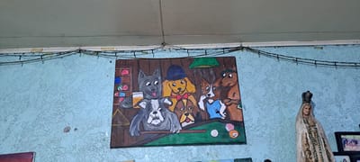 The media file shows a painting of several anthropomorphic dogs playing billiards. The dogs are depicted with human-like expressions and clothing, including a grey dog wearing a bowler hat and a tan dog with a red bowtie. They are gathered around a green pool table, with some holding cues and others looking at the game. There are also pool balls and a rack visible in the painting. The painting itself is mounted on a light blue, textured wall. Above the painting, a string of small lights is strung across the wall. To the right of the painting, a statue of the Virgin Mary wearing a crown and holding a rosary is visible. There are also framed photographs in the background. The overall setting appears to be indoors, possibly in a home or a casual establishment. The style of the painting is whimsical and cartoonish.