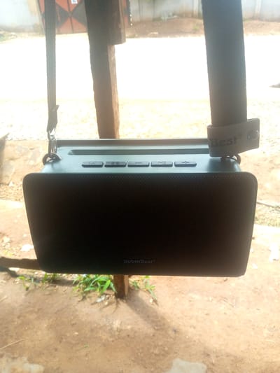 An outdoor scene in Madakiya, Nigeria, features a dark green, rectangular portable Bluetooth speaker hanging from a strap attached to a wooden post. The speaker has a textured front grille with the brand name "BoomBest" subtly visible on the lower right side. The top surface of the speaker has several buttons, including "M" for mode, "EQ" for equalizer, and volume control buttons represented by "-" and "+". A leather strap with the embossed word "Best" and a small circle is attached to the speaker via a metal clasp. The background shows blurred foliage, soil, and what appears to be a structure with an orange and black pattern. The lighting suggests it is daytime.