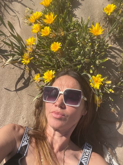 A person lies on a sandy beach next to a clump of yellow flowers. They are wearing sunglasses and a black and white shoulder strap with the word "Armani" printed on it. The location appears to be La Marina De Creixell, Spain, given the coastal environment, sandy terrain, and possible vegetation. The image is taken from an overhead perspective.