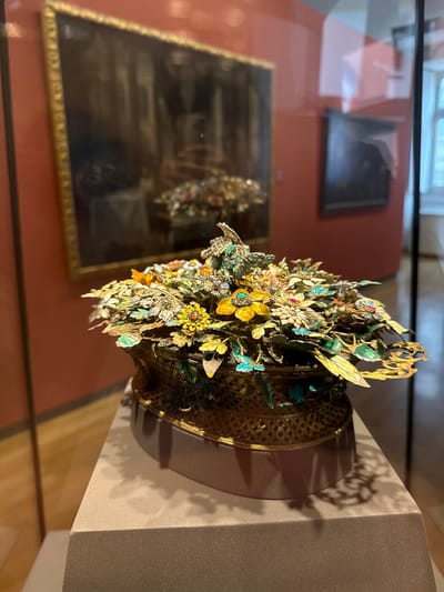 The image shows a floral crown in a museum display case in Biberach an der Riß, Germany. The crown is made of gold and features various flowers and leaves. It sits on a grey display stand. In the background, reflections show an artwork, possibly a painting, and the museum's interior. The lighting is a mix of natural light and artificial illumination, highlighting the intricate details of the crown.