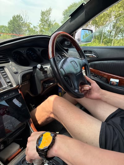 The image shows the interior of a car, with a person's hands on the steering wheel and gear shift. The driver is wearing a large, dark watch with a yellow and blue face. The car's dashboard is visible, including the speedometer and other gauges. The exterior of the car, with trees and a cloudy sky, can be seen through the windows. The scene appears to be set in Chernigovka, Russia, based on contextual information.