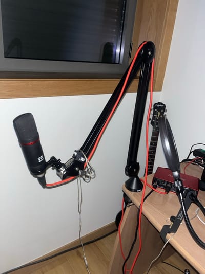 This image depicts an indoor scene in Pedrouços, Portugal, primarily featuring a recording setup. A black RØDE microphone is mounted on a black RØDE boom arm, which is clamped to a light-colored wooden desk situated against a white wall. Red and black cables are routed along the boom arm and across the desk. On the desk, a red Focusrite Scarlett 2i2 audio interface is visible with multiple cables connected. A black pop filter, attached to a flexible arm, is also clamped to the desk. Behind the audio interface and pop filter, the headstock and upper neck of a dark-colored Epiphone electric guitar are visible, leaning against the white wall. A window with horizontal blinds is present in the upper left, featuring a light wood frame. The floor is covered with light wood flooring. No individuals are present in the scene.