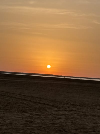 A sunset is depicted over the beach and the Mediterranean Sea in Port-la-Nouvelle, France. The sun, a bright orange orb, hangs low in the sky, casting a warm glow across the scene. The sky transitions from a deep orange near the horizon to a lighter, peachy hue overhead, with subtle streaks of clouds. The foreground is dominated by the sandy beach, with visible tire tracks creating patterns in the sand. The sea is a dark, reflective strip separating the beach from the distant horizon. Two small figures, likely people, are visible in the distance on the beach, adding a sense of scale to the vastness of the landscape. The overall impression is one of tranquility and natural beauty.
