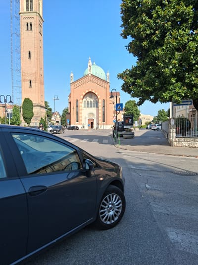 A dark-colored car is parked on the right side of a street in what appears to be a town or city. The car is partially obscuring a view of a large, historic brick building with a prominent green dome, possibly a church or cathedral, on the left side of the frame. Next to this building, a tall brick tower stands. The sky is a clear, bright blue, suggesting a sunny day. Trees with lush green foliage are visible, one large tree in the upper left corner and smaller trees and bushes scattered throughout the scene.

In the background, other buildings and more trees can be seen lining the street. Traffic lights and street signs are also visible. One sign in the distance has text that appears to be "Alzate" and another sign closer to the foreground reads "Santimello". The scene suggests an urban environment with historical architecture. The time of day is likely daytime, given the bright sunlight and clear blue sky. There are no people prominently featured in the image, but the presence of a car and parked vehicles indicates daily life. The overall mood is calm and serene, capturing a moment of stillness in a European town.