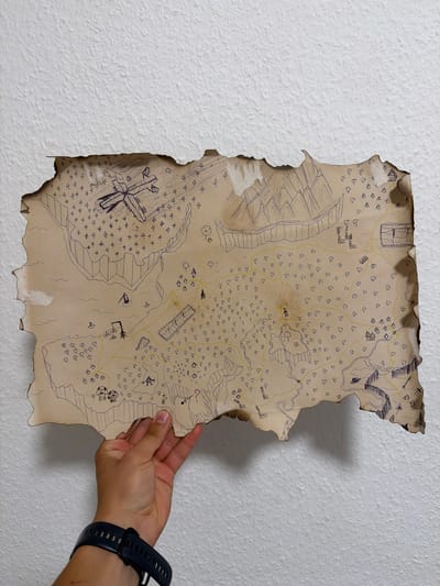 A hand is holding a burnt, torn, hand-drawn map against a textured white wall. The map depicts a fantastical landscape with various drawn elements including mountains, trees, bodies of water, buildings, a bridge, a possibly crashed aircraft, and a question mark. The map is drawn in dark ink on aged-looking paper, with the edges appearing charred and irregular. The hand holding the map is positioned at the bottom left of the frame, with a dark watch on the wrist. The background is a plain white wall with a rough texture. There is no discernible connection to the city of Nienburg/Weser, Germany in this image.