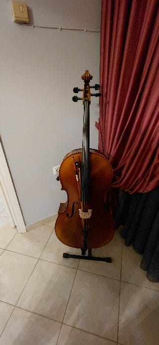 An indoor scene, situated in the Municipal Unit of Acharnes, Greece, displays a dark brown cello positioned upright on a black metal floor stand. A wooden bow rests vertically against the cello's strings and body. The cello's headstock with tuning pegs is visible at the top. The background consists of a light grey wall on the left, featuring a beige rectangular light switch with a "1" symbol and a white electrical conduit running horizontally below it. To the right, pleated red curtains hang, with a section of dark, possibly black, fabric visible beneath them. The floor is tiled with light-colored square tiles. No individuals are present, and no actions or interactions are depicted.