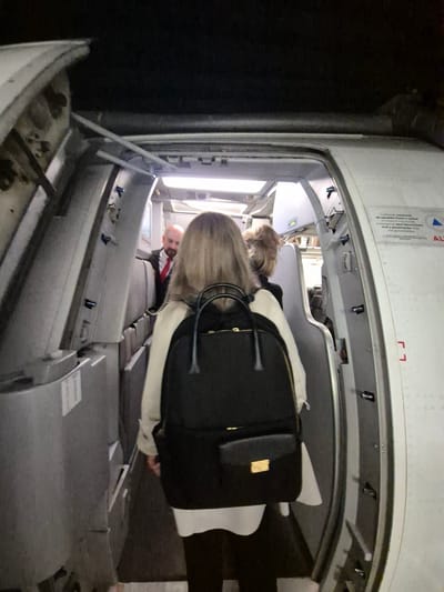 The image shows the interior of what appears to be an airplane. A woman with blonde hair is bent over, possibly placing luggage in an overhead compartment. Another person, possibly a male, wearing a red tie and a black suit can be seen in the background. The overhead compartment is open, revealing the interior with lighting and some seated passengers further down the aisle. The area outside the compartment appears to be dimly lit or dark. The photograph also contains some text related to conduct guidelines, potentially indicating safety or passenger regulations on the plane. The location in Rionegro, Colombia is implied, as the safety guidelines are in Spanish.