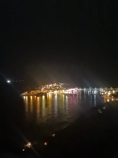 The image displays a nocturnal aerial or elevated view of the settlement of Husøy, Norway, situated on a landmass surrounded by water. The sky is dark, indicating night conditions. Numerous artificial lights from buildings and streetlights illuminate the town, appearing predominantly yellow and orange, with some red and blue lights visible, particularly along the waterfront. These lights are reflected distinctly on the calm water surface below, creating elongated streaks of color.
The town's lights ascend the terrain, suggesting it is built on a hillside or elevated area, with a prominent red light visible on a structure at a higher elevation within the settlement. To the right, a multi-span bridge, marked by regularly spaced white lights, extends across a section of the water, connecting to the main landmass or another part of the island. Scattered, smaller light sources are present on the water, including a distinct bright white light in the upper left section of the water and additional fainter lights in the distance, possibly from boats or distant shorelines. No individuals or specific actions are discernible within the frame.