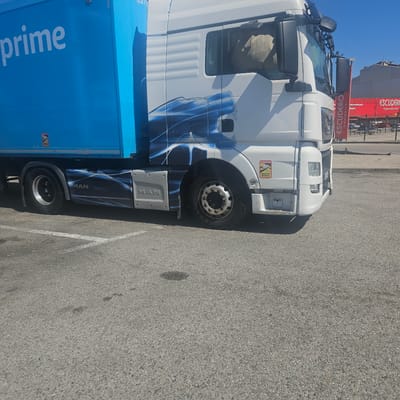A white and blue Amazon Prime delivery truck is parked in a parking lot in La Jonquera, Spain. The truck has an MAN logo on the side and "prime" printed in large white letters on the blue cargo box. The truck is parked on a grey asphalt surface, which is marked with a white parking line. In the background, a red building with the sign "ESCUDERO hipermercat" is visible, indicating the location in Spain. The sky is blue and clear. There are "Attention Angles Morts" stickers on both the cab and cargo box of the truck.