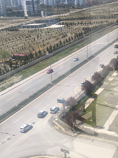 The image provides an elevated, oblique view of a multi-lane road and an adjacent urban landscape under bright daylight. The setting is likely Etimesgut, Turkey, as indicated by a prominent sign.

In the foreground, a wide, multi-lane asphalt road dominates, featuring white lane markings, including dashed lines, solid lines, and crosswalks. Several cars are visible: a red sedan and a white hatchback occupy the left-hand lanes, while a white sedan and a silver hatchback are further down on the right-hand side. Closer to the bottom, two white hatchbacks with distinct markings (possibly taxis or service vehicles) and a gray hatchback are situated near an intersection or turn-off. The road is flanked by grassy medians containing low bushes, concrete barriers, and patterned sidewalks. Streetlights are spaced along the roadside.

Beyond the road, a large expanse of land is visible, characterized by numerous young, conifer-like trees planted in orderly rows, suggesting a recent landscaping or development project. In this area, several modern, flat-roofed buildings appear to be new or under construction. In the distant background, a dense concentration of modern, multi-story apartment buildings rises, indicating a residential or commercial urban center.

Notable details include significant reflections of geometric patterns on the window through which the photograph was captured. A blue sign near the bottom right clearly displays the text "DÜNYA VADİSİ BAŞKANLIĞI," which translates to "World Valley Presidency," likely referring to a residential or municipal development. The sky is clear, and the bright sunlight casts subtle shadows, suggesting midday or early afternoon. No individuals are explicitly visible, though the presence of vehicles implies drivers.