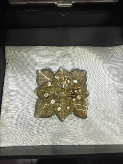 The scene depicts a single, decorative metallic object, resembling a brooch or buckle, centered within an open dark-colored box, presumed to be a jewelry case, in the city of Liège, Belgium. The object is approximately 5-6 cm in its widest dimension, crafted from an aged, possibly tarnished brass or bronze-toned metal, exhibiting areas of greenish patina and darker discoloration. It is designed as a stylized flower or cluster of leaves with a symmetrical, roughly square outline. The design features interconnected petals or leaves, each intricately detailed with an openwork or filigree pattern mimicking venation. Embedded within the metallic structure are approximately 18-20 small, round, faceted clear stones, likely rhinestones, arranged in clusters of two to four on the individual elements of the design. The object rests on a light silver-grey, subtly textured fabric lining, possibly satin or silk, which forms the bottom interior of the box. The interior sides of the box are dark, appearing black with a matte finish. A metallic hinge or clasp component is partially visible along the top interior edge of the box. No people or actions are visible.