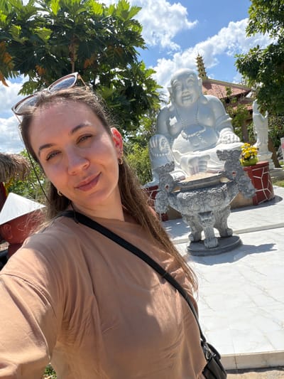 A woman is taking a selfie outdoors in what appears to be a temple complex. She is wearing a tan t-shirt and sunglasses on her head. Behind her is a large white statue of a laughing Buddha, with a small incense burner in front of it. In the background, there are trees, a tiled roof building, and a pagoda. The ground is paved with white tiles. The scene suggests the location is in Tuy An Nam, Vietnam, a region known for its temples and pagodas.