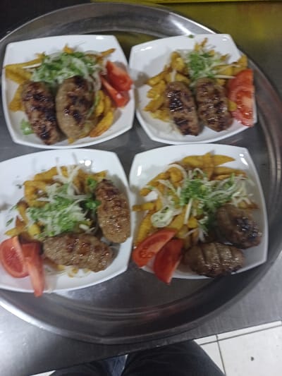 Four square white plates arranged on a large metal tray hold identical portions of food. Each plate contains two grilled meat rolls alongside a bed of french fries, topped with shredded onions and fresh greens. A slice of tomato accompanies the meal. The arrangement of the plates on the tray creates a symmetrical pattern.